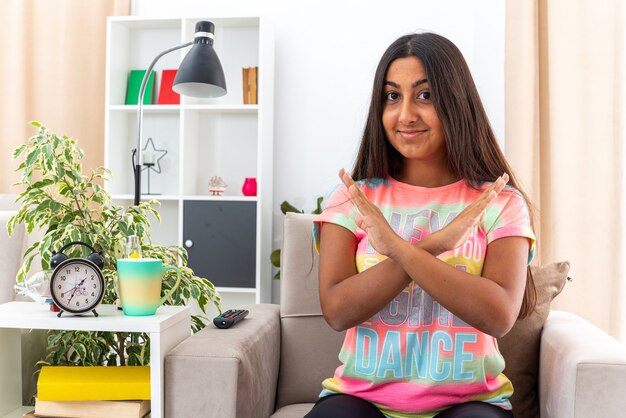 Junges Mädchen in Freizeitkleidung, das mit einem Lächeln im Gesicht schaut und eine Stoppgeste macht, die die Hände kreuzt, die auf dem Stuhl im hellen Wohnzimmer sitzen?