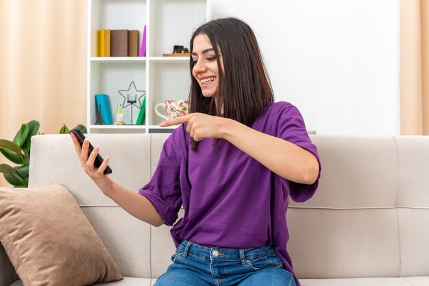 Junges Mädchen in Freizeitkleidung, das Smartphone hält, das mit dem Zeigefinger darauf zeigt, glücklich und fröhlich lächelt breit auf einer Couch im hellen Wohnzimmer sitzend