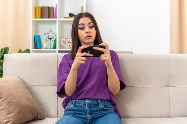 Junges Mädchen in Freizeitkleidung, das Spiele mit Smartphone spielt und selbstbewusst auf einer Couch im hellen Wohnzimmer sitzt