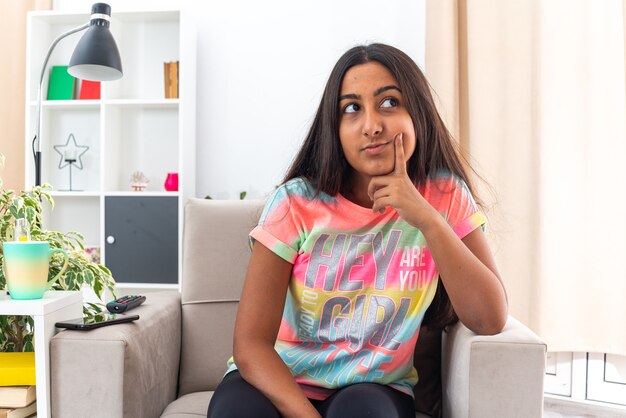 Junges Mädchen in Freizeitkleidung, das verwirrt auf dem Stuhl im hellen Wohnzimmer sitzt