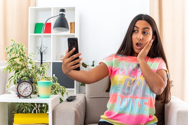 Junges Mädchen in Freizeitkleidung macht Selfie mit Smartphone und schaut erstaunt und überrascht auf dem Stuhl im hellen Wohnzimmer sitzend