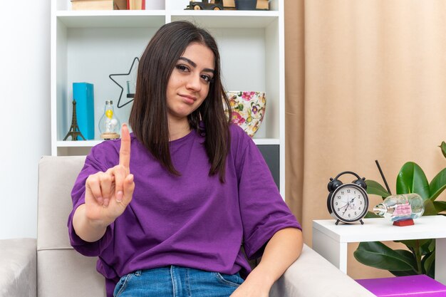 Junges Mädchen in Freizeitkleidung mit ernstem Gesicht, das Zeigefinger mit skeptischem Ausdruck auf einem Stuhl im hellen Wohnzimmer zeigt