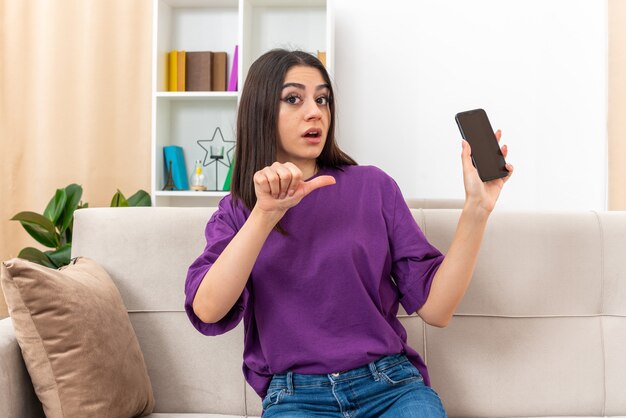Junges Mädchen in Freizeitkleidung mit Smartphone, das verwirrt aussieht und mit dem Daumen auf das Handy zeigt, das auf einer Couch im hellen Wohnzimmer sitzt?