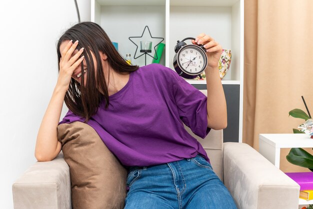 Junges Mädchen in Freizeitkleidung mit Wecker, das fröhlich lächelnd das Gesicht mit der Palme bedeckt, die auf einem Stuhl im hellen Wohnzimmer sitzt sitting