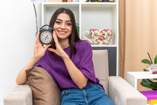 Junges Mädchen in Freizeitkleidung mit Wecker sieht glücklich und positiv aus und lächelt fröhlich auf einem Stuhl im hellen Wohnzimmer sitzen