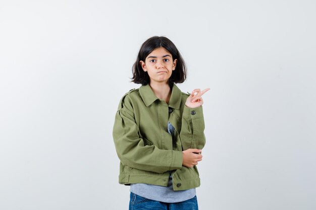 Junges Mädchen in grauem Pullover, Khaki-Jacke, Jeanshose, die nach rechts zeigt, während sie die Hand am Ellbogen hält und süß aussieht, Vorderansicht.