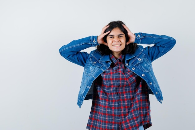 Junges Mädchen in kariertem Hemd und Jeansjacke, das die Hände auf den Kopf hält und glücklich aussieht, Vorderansicht.