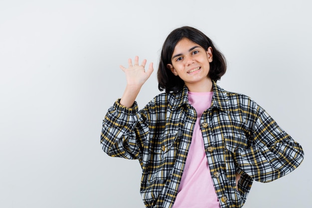 Junges Mädchen in kariertem Hemd und rosa T-Shirt, das die Hand ausstreckt, um jemanden zu begrüßen, während es die Hand an der Taille hält und süß aussieht looking