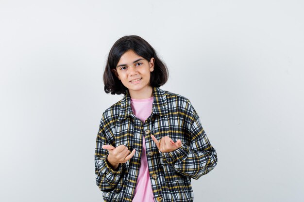 Junges Mädchen in kariertem Hemd und rosa T-Shirt, das kleine Finger und Rock'n'Roll-Geste zeigt und süß aussieht, Vorderansicht.