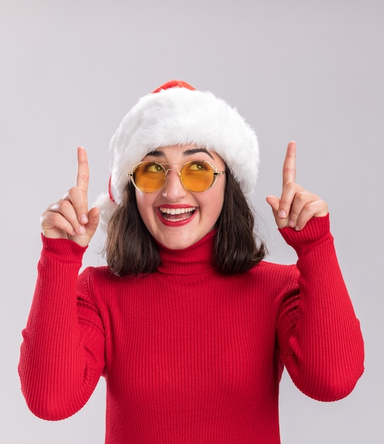 Kostenloses Foto junges mädchen in rotem pullover und weihnachtsmütze mit brille, das glücklich und freudig aufschaut und mit den zeigefingern nach oben auf weißem hintergrund zeigt