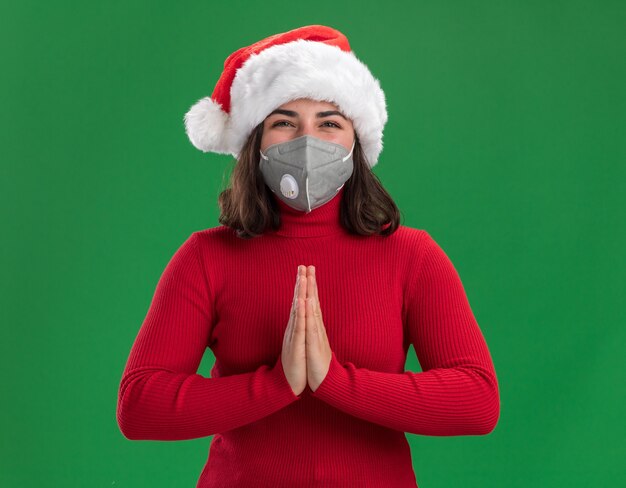 Junges Mädchen in rotem Pullover und Weihnachtsmütze tragen Gesichtsschutzmaske mit glücklichem Gesicht, das Hände zusammenhält wie namaste Geste, die über grüner Wand steht