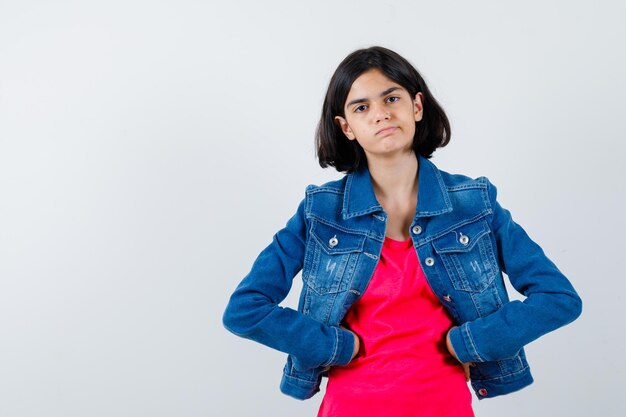 Junges Mädchen in rotem T-Shirt und Jeansjacke, das die Hände an der Taille hält und ernst aussieht, Vorderansicht.