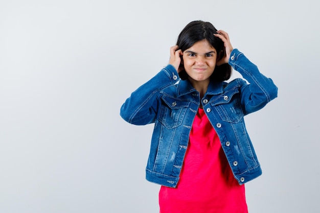 Junges Mädchen in rotem T-Shirt und Jeansjacke, das die Hände auf den Kopf drückt und glücklich aussieht, Vorderansicht.