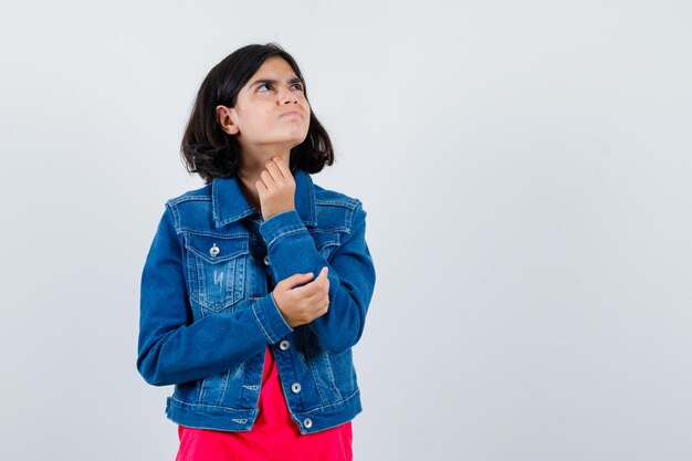 Junges Mädchen in rotem T-Shirt und Jeansjacke, das die Hand am Ellbogen hält und nach oben schaut und nachdenklich aussieht, Vorderansicht.