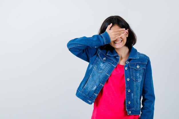 Junges Mädchen in rotem T-Shirt und Jeansjacke, die die Augen mit der Hand bedeckt und süß aussieht, Vorderansicht.