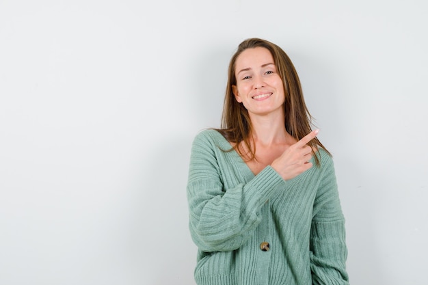 Junges Mädchen in Strickwaren, die rechts mit dem Zeigefinger zeigen und glücklich schauen, Vorderansicht.