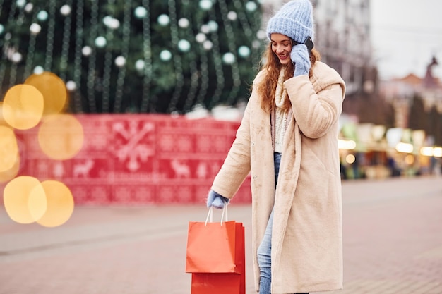Junges mädchen in warmer kleidung mit einkaufstüten in der hand macht einen spaziergang im freien in der stadt und hält das telefon in der hand.