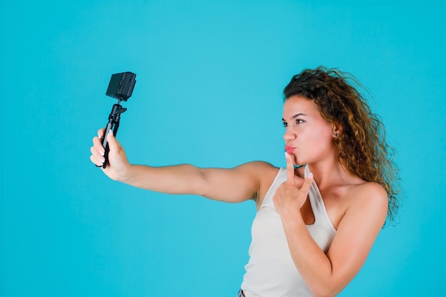 Junges Mädchen macht Selfie mit ihrer Mini-Canera, indem sie den Zeigefinger auf den Lippen auf blauem Hintergrund hält