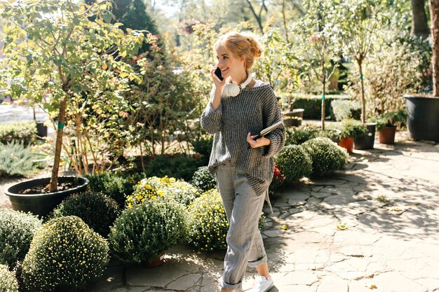 Junges Mädchen mit Brötchen geht im botanischen Garten entlang der Gasse und telefoniert Porträt einer hübschen Frau mit Kopfhörern um den Hals und Notizbuch in den Händen