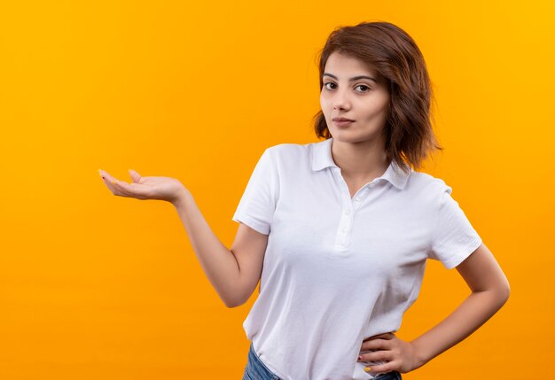 Junges Mädchen mit dem kurzen Haar, das Poloshirt trägt, das zuversichtlich mit Arm der Hand präsentiert