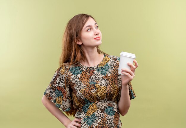 Junges Mädchen mit dem langen Haar, das buntes Kleid hält Kaffeetasse hält, das zuversichtlich schaut beiseite schaut