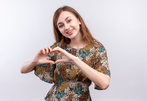 Junges Mädchen mit dem langen Haar, das buntes Kleid positiv und glücklich lächelnd breit tut, macht Herzgeste mit Hand über Brust