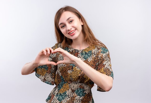 Junges Mädchen mit dem langen Haar, das buntes Kleid positiv und glücklich lächelnd breit tut, macht Herzgeste mit Hand über Brust