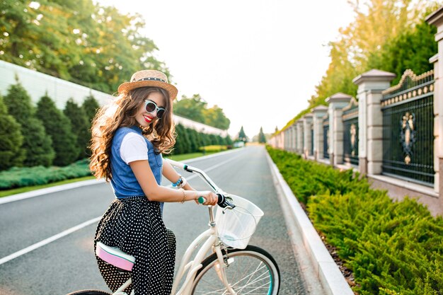 Junges Mädchen mit dem langen lockigen Haar in der blauen Sonnenbrille fährt mit dem Fahrrad auf der Straße. Sie trägt einen langen Rock, ein Wams und einen Hut. Sie lächelt. Blick von hinten.
