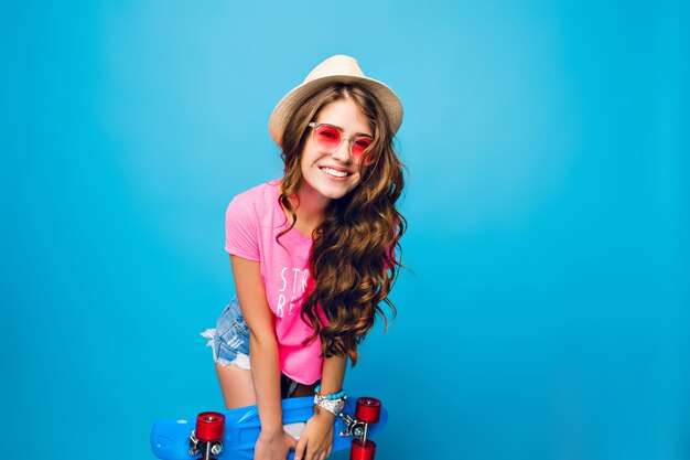 Junges Mädchen mit dem langen lockigen Haar in der rosa Sonnenbrille, die auf blauem Hintergrund im Studio aufwirft. Sie trägt Shorts, rosa T-Shirt, Hut. Sie hält ein blaues Skateboard und lächelt in die Kamera.