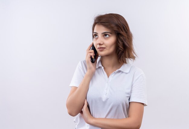 Junges Mädchen mit den kurzen Haaren, die weißes Poloshirt tragen, das zuversichtlich spricht, während auf Handy spricht