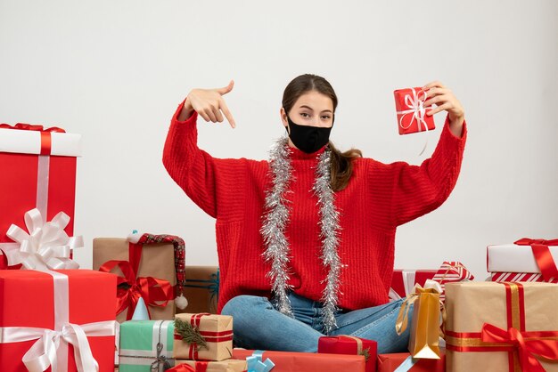 junges Mädchen mit der schwarzen Maske, die Geschenk hält, das auf sich selbst zeigt, das um Geschenke auf Weiß sitzt