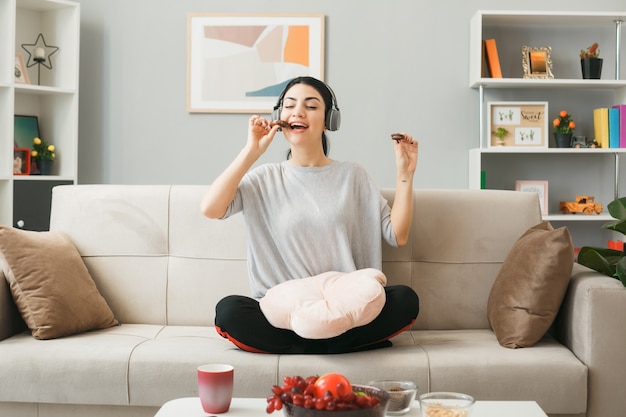 Junges Mädchen mit Kissen, das Kopfhörer trägt, isst Kekse, die auf dem Sofa hinter dem Couchtisch im Wohnzimmer sitzen?