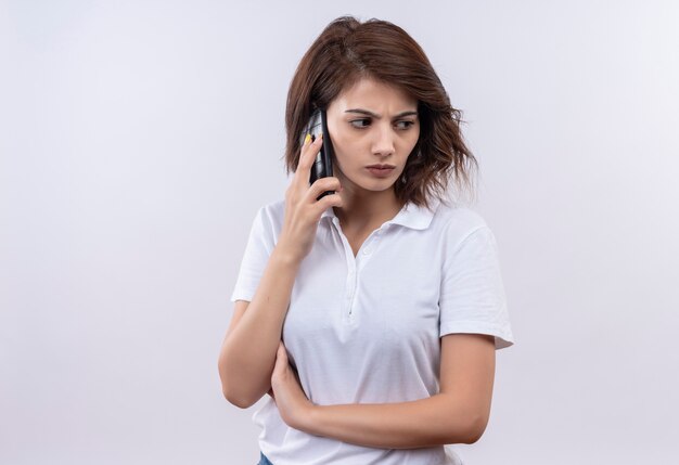 Junges Mädchen mit kurzen Haaren, die weißes Poloshirt tragen, das auf Handy mit stirnrunzelndem Gesicht spricht