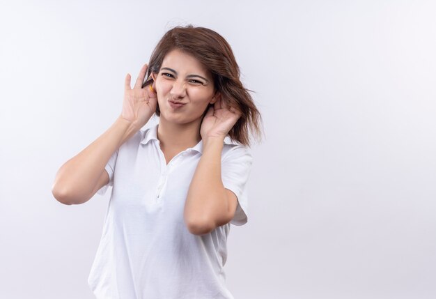 Junges Mädchen mit kurzen Haaren, die weißes Poloshirt tragen, das Grimasse macht, das große Ohren mit Händen nachahmt