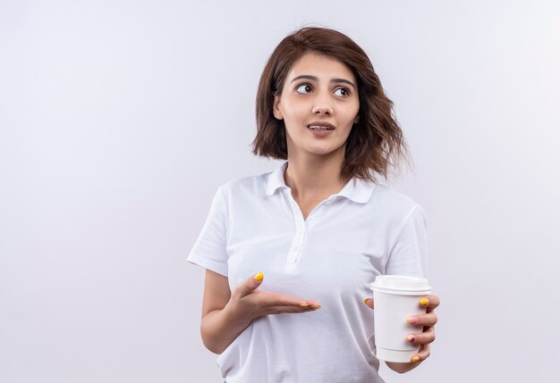 Junges Mädchen mit kurzen Haaren, die weißes Poloshirt tragen, das Kaffeetasse präsentiert, die verwirrt schaut