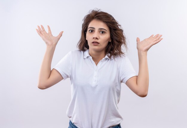 Junges Mädchen mit kurzen Haaren, die weißes Poloshirt tragen, das unsicher und verwirrte Achselzucken schaut und keine Antwort hat