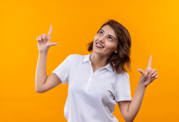 Junges Mädchen mit kurzen Haaren, die weißes Poloshirt tragen und fröhlich mit Zeigefingern nach oben zeigen