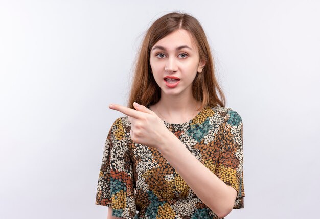 Junges Mädchen mit langen Haaren, die buntes Kleid tragen, das verwirrt zeigt und mit Zeigefinger zur Seite zeigt