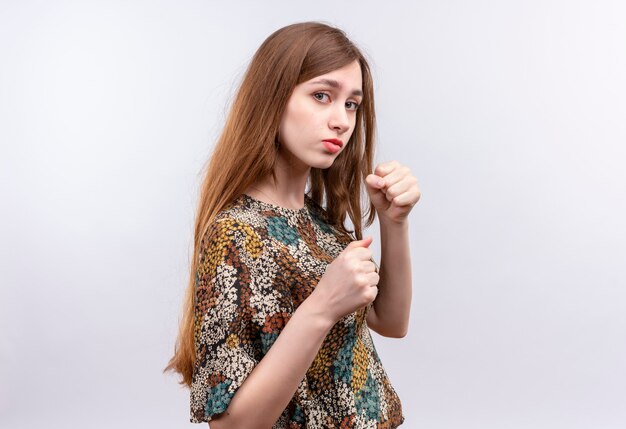 Junges Mädchen mit langen Haaren, die buntes Kleid tragen Kamera mit ernstem Gesicht betrachten, das wie ein Boxer mit geballten Fäusten aufwirft