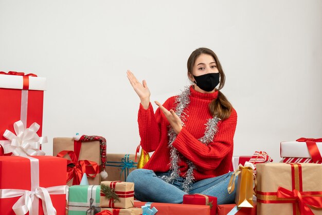 junges Mädchen mit rotem Pullover und schwarzer Maske, die auf etwas zeigt, das um Geschenke auf Weiß sitzt