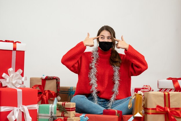 junges Mädchen mit rotem Pullover und schwarzer Maske, die Fingerpistolen zu ihrer Schläfe setzen, die um Geschenke auf Weiß sitzen