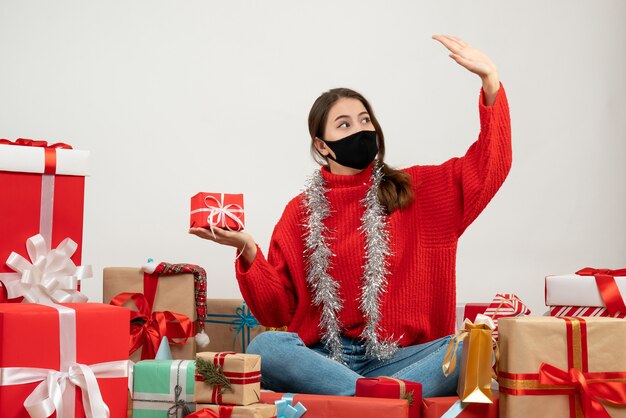 junges Mädchen mit rotem Pullover und schwarzer Maske, die Geschenk hält, das ihre Hand hebt, die um Geschenke auf Weiß sitzt