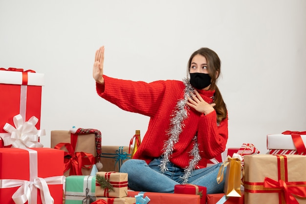 junges Mädchen mit rotem Pullover und schwarzer Maske, die Hand auf ihre Brust setzt, die Geschenke auf Weiß sitzt