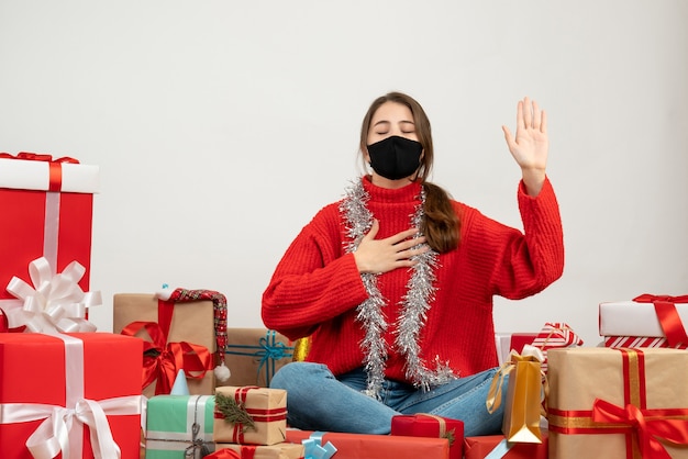 junges Mädchen mit rotem Pullover und schwarzer Maske, die Versprechenszeichen macht, das um Geschenke auf Weiß sitzt