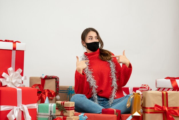junges Mädchen mit rotem Pullover und schwarzer Maske zeigt auf sich selbst sitzend um Geschenke auf Weiß