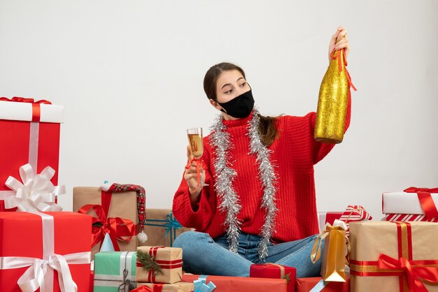 junges Mädchen mit schwarzer Maske, die Champagner hält, der Geschenke auf Weiß sitzt