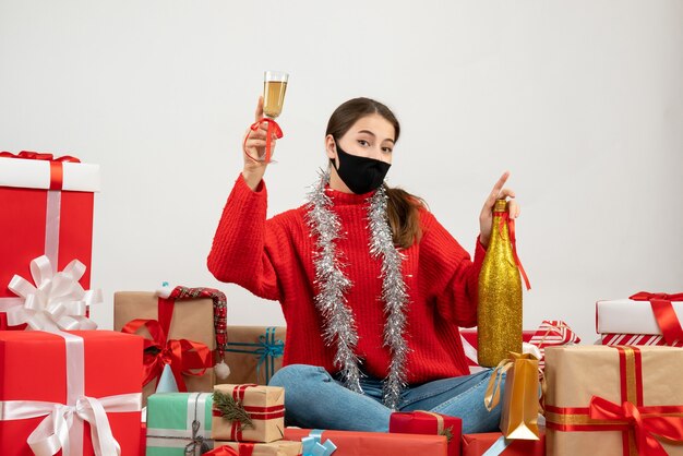 junges Mädchen mit schwarzer Maske, die Champagnerflasche hält und röstend sitzt Geschenke auf Weiß