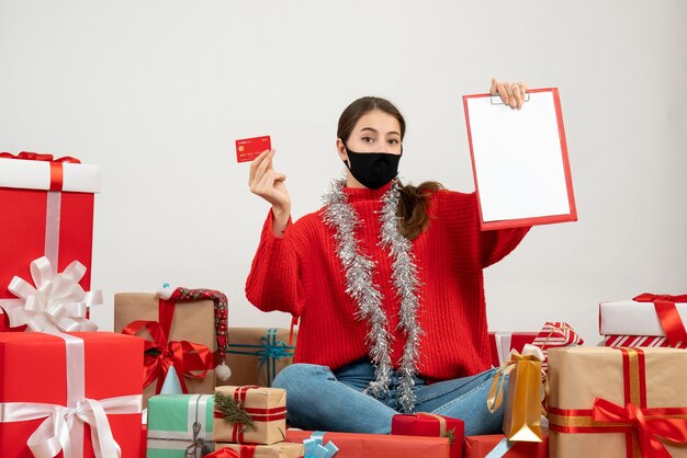 junges Mädchen mit schwarzer Maske, die Karte und Dokument hält, das um Geschenke auf Weiß sitzt