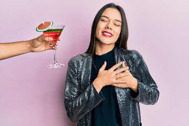 Junges Mädchen mit sexy Partyjacke und hispanischem Arm, das einen Cocktail anbietet, lächelt mit den Händen auf der Brust, die Augen geschlossen und eine dankbare Geste im Gesicht. Gesundheitskonzept.