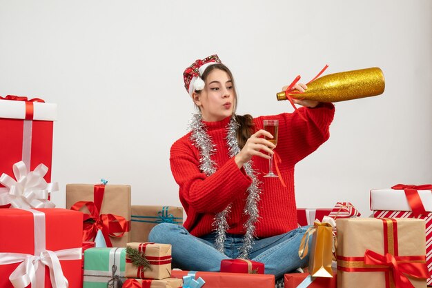 junges Mädchen mit Weihnachtsmütze, die Champagner gießt, der um Geschenke auf Weiß sitzt